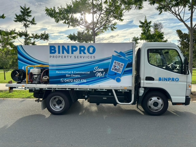 The image shows a corrugated metal roof, angled downward, with a background of an asphalt area. The roof panels are light gray, and the surface appears clean and well-maintained, illuminated by sunlight.