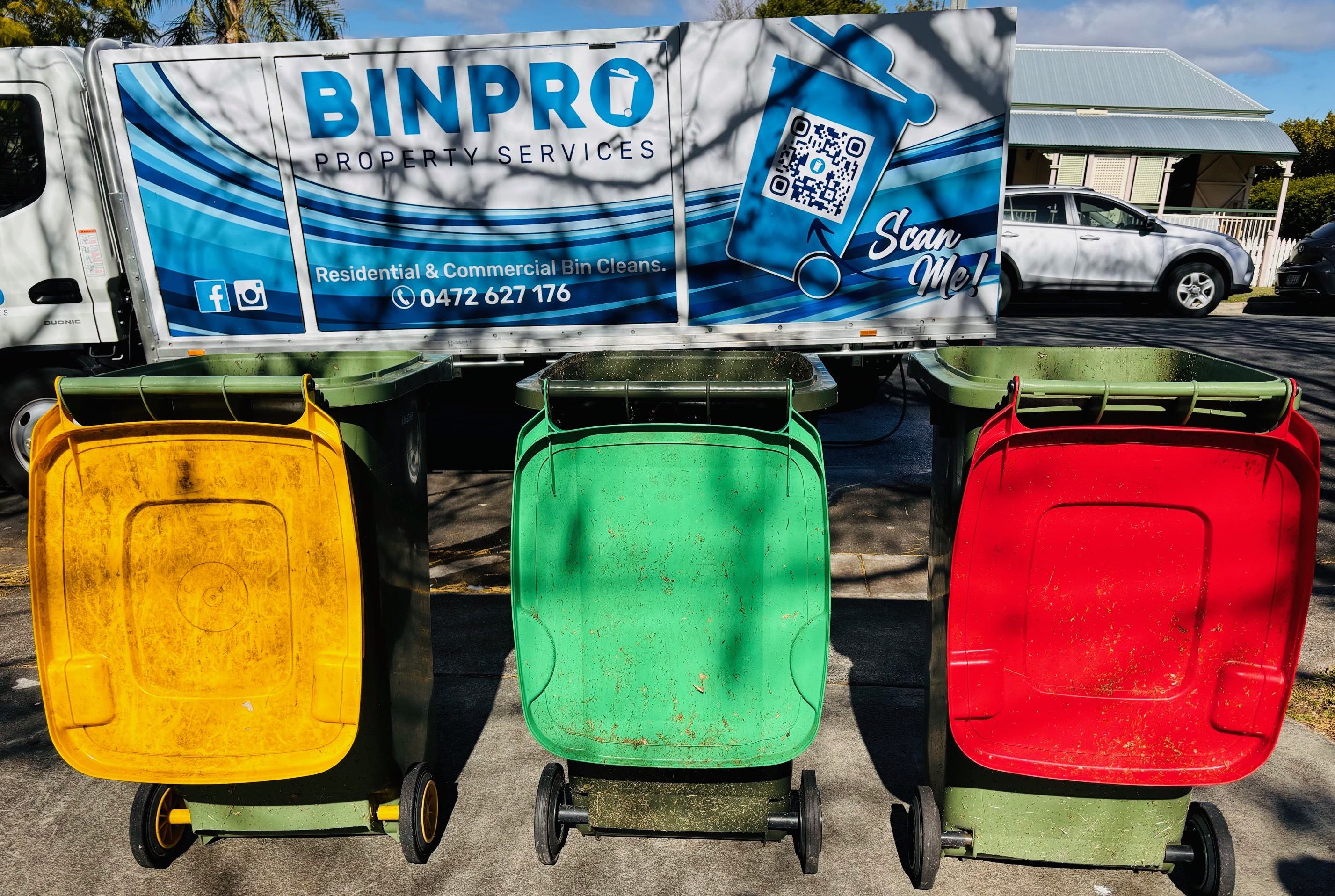 A person in a white cap and shorts is cleaning solar panels on a rooftop using a long brush and hose. The building has light-colored walls and dark-framed windows. The sky is clear and blue.